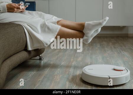Woman Relaxing on sofa. Automatic robotic vacuum cleaner on laminate wood floor, smart cleaning technology Stock Photo