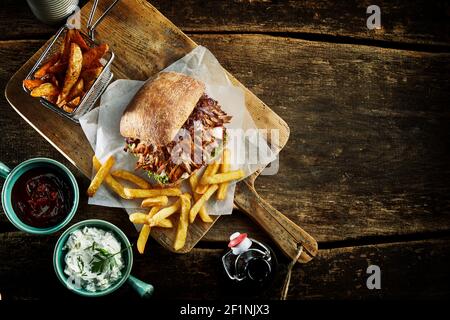 Top view of delicious pulled pork burger and sauces placed on wooden table with rustic potatoes and French fries in cafe Stock Photo