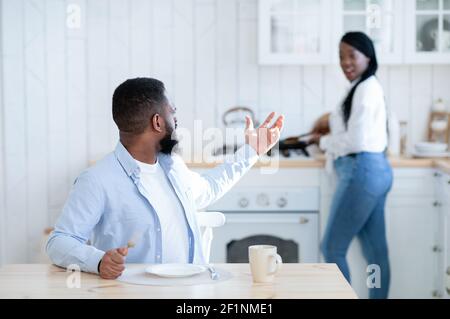 Hungry african husband complaining about empty plate, waiting for dinner in kitchen while his spouse cooking food on background, starving black man si Stock Photo