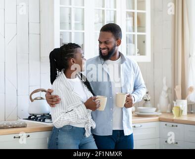 Romantic African American Spouses Drinking Coffee And Chatting In Kitchen. Cheerful Black Millennial Couple Having Hot Caffeine Drinks And Enjoying Sp Stock Photo