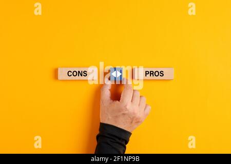 Male hand holds a wooden cube with arrow icon between the options of pros or cons. The decision between the pros and cons of a situation.. Stock Photo