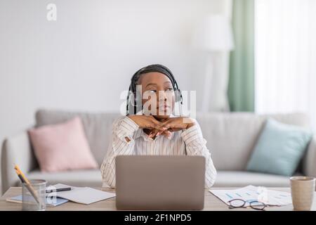 Pensive young black lady having online training at home, sitting at workdesk in front of laptop, leaning on her elbows and looking at copy space, usin Stock Photo
