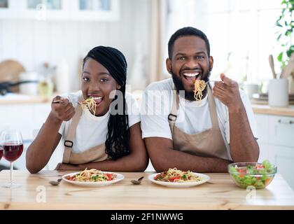 Cheerful Black Spouses Sitting At Table Eating Tasty Lunch In Kitchen, Happy African American Couple Having Spaghetti And Vegetable Salad, Enjoying De Stock Photo