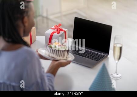 Closeup of unrecognizable african american woman holding birthday cake, using laptop with blank screen, sitting at desk with gift box and glass of cha Stock Photo
