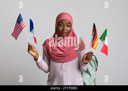 Confused african american woman in hijab showing diverse flags, carrying backpack on shoulder, grey background. Muslim black lady student with bunch o Stock Photo