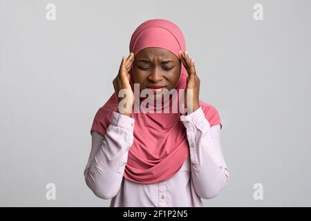 Upset young muslim african american woman in hijab having headache, touching her head, closing eyes, grey studio background, copy space. Migraine, hea Stock Photo