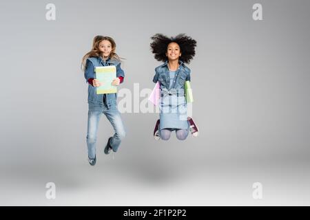 cheerful interracial schoolkids in stylish denim clothes jumping with books on grey Stock Photo