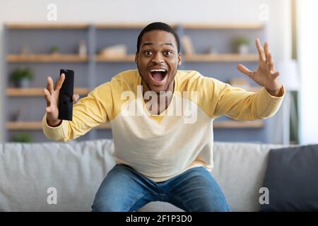 Happy african american man having fun at home, screaming with remote controller in his hand, watching football game on TV, empty space. Black guy enjo Stock Photo