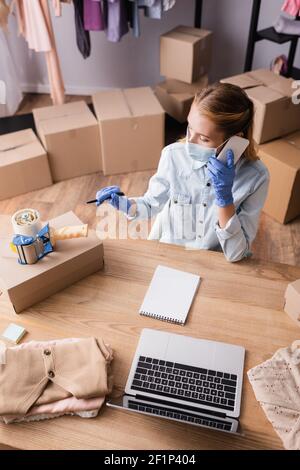 high angle view of showroom owner in medical mask, talking on smartphone near laptop Stock Photo