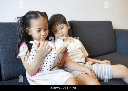 Preteen Vietnamese brother and sister sitting on sofa and reading book of stories together Stock Photo