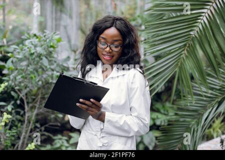 Plant care and protection concept. Work in the greenhouse. Pleasant smiling african american female agricultural engineer stands in front of beautiful Stock Photo