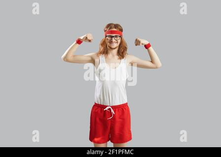 Funny skinny man in tank top and shorts showing weak muscles isolated on gray background Stock Photo