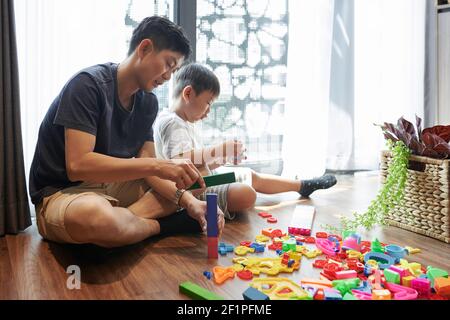 Father and preteen son sitting on the floor at home and building towers with plastic blocks Stock Photo