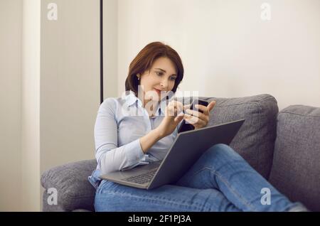 Attractive middle age woman with laptop and smartphone sitting indoors on sofa Stock Photo