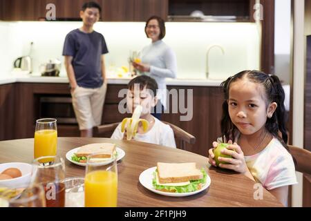Preteen Vietnamese children sitting at kitchen table and eating sandwiches and fruits for breakfast Stock Photo