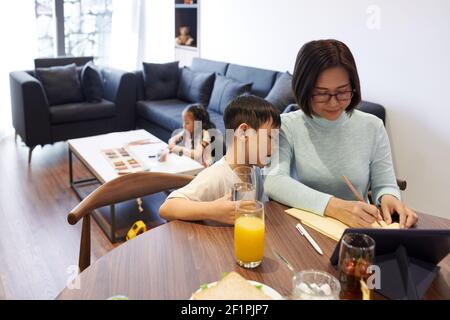 Curious small boy drinking glass of water and looking at his mother writing shopping list or plas for the day Stock Photo
