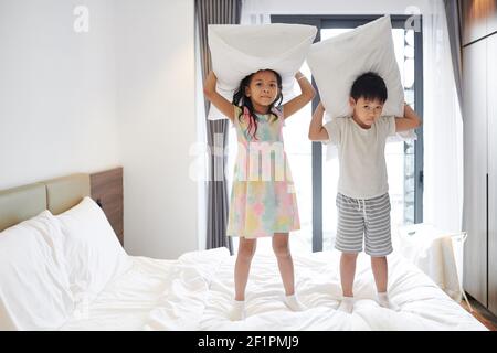 Vietnamese brother and sister playing with pillows on bed in parents bedroom and looking at camera Stock Photo