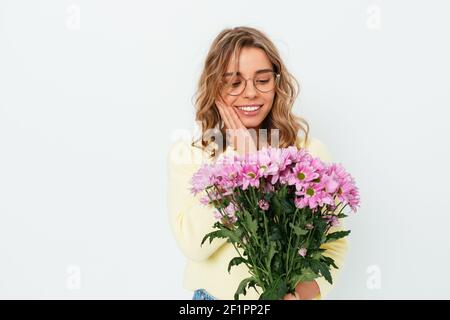 Beautiful happy young blonde woman wearing eyeglasses and casual outfit holding looking at bouquet pink flowers standing over white studio background, Stock Photo