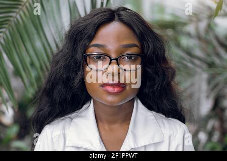 Close up portrait of young African woman, scientist, botanist, agronomist, wearing glasses and white lab coat, posing to camera over the palm trees on Stock Photo