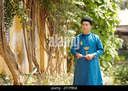 Handsome young Vietnamese man in ao dai dress walking on street with paper fan in hands Stock Photo