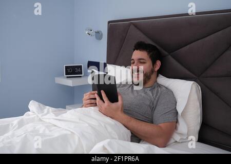 Young latin man reading a book and smiling in bed late in the morning. Digital clock at the background. Stock Photo