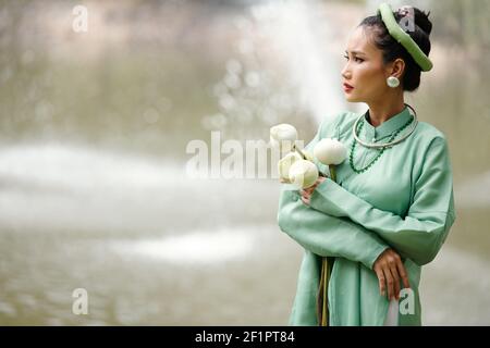 Worried gorgeous young Vietnamese woman in traditional costume standing by river with lotus flowers in hands and looking at water Stock Photo
