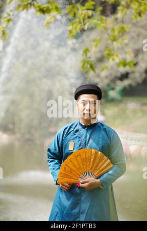 Portrait of handsome young Vietnamese man in ao dai dress and traditional headpiece holding paper fan and smiling at camera Stock Photo