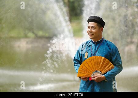 Happy young Asian man in traditional Vietnamese costume and headpiece standing by river with paper fan in hand and looking away Stock Photo