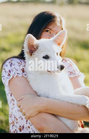 Stylish happy woman hugging cute white puppy in warm sunset light in summer meadow. Casual young female holding adorable swiss shepherd fluffy puppy. Stock Photo