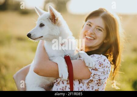Stylish happy woman hugging cute white puppy in warm sunset light in summer meadow. Casual young female holding adorable swiss shepherd fluffy puppy. Stock Photo