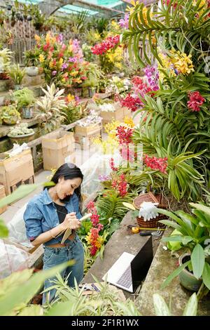 Positive female greenhouse worker talking on phone with customer and taking notes in planner Stock Photo