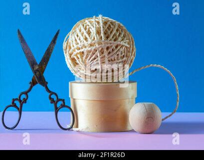 ball of string on a wooden pedestal with a pair of ornate scissors on a blue and pink background , concept of hobbys  and pastimes . Stock Photo