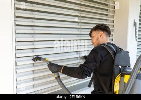 Professional cleaner vacuum cleaning window blinds on an apartment balcony in a high-rise building w Stock Photo