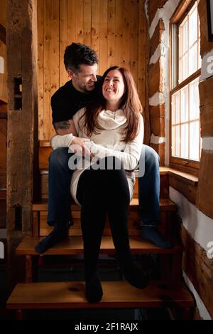 Happy couple sitting on stairs together inside rustic log cabin. Stock Photo