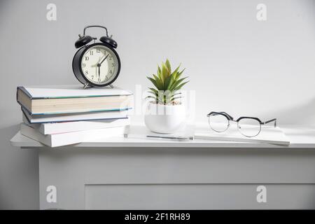 A stack of books, an artificial succulent plant in a ceramic pot, glasses, a metal basket with notebooks, a pen are on a white table surface. Concept Stock Photo