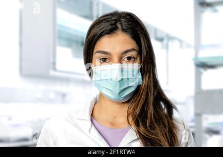A brunette doctor with a surgical mask with a blurred medical laboratory in the background. Perfect shot for pandemic, research, vaccine and security. Stock Photo