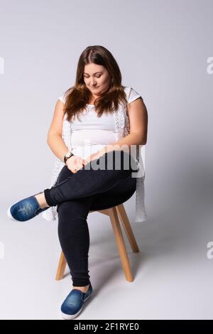 An overweight young woman sits on a chair and looks thoughtfully at her body. Photo in full growth on a gray background. Stock Photo