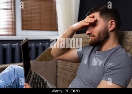 Profile photo of a tired caucasian business man sitting at home on the couch with a laptop. Work from home online. Stock Photo