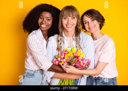 Photo of nice optimistic three ladies hug hold flowers wear white shirt isolated on bright yellow color background Stock Photo