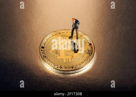 Businessman miniature figure standing on a gold Bitcoin looking down thinking about the risks and returns of investing in the cryptocurrency Stock Photo