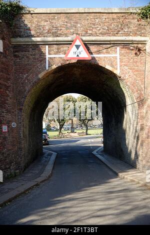 Railway bridge over a road in England. Triangular warning road sign depicts maximum height. Brick arch with a green park on the far side. Stock Photo