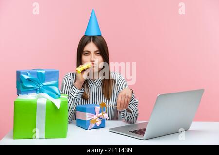 Unhappy brunette woman with party cone and blower sitting at workplace surrounded by many presents, feeling depressed celebrating birthday at work. In Stock Photo