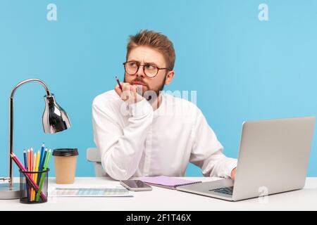 Thoughtful bearded man journalist working on article siting at workplace with laptop using his notes in notepad, thinking about headline. Indoor studi Stock Photo