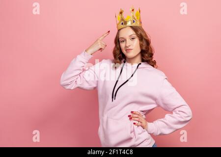 Portrait of ambitious curly haired teenage girl in hoodie pointing finger on crown, feeling princess, confidence. Indoor studio shot isolated on pink Stock Photo
