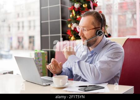A middle-aged caucasian man with a headset in eyewear talking via video link with smile. He works during a coffee break with a laptop, mobile phone and papers on the table sitting in a cafe. SDOF Stock Photo