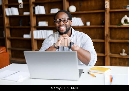 Cheerful African-American guy in smart casual wear sits at the desk with a laptop, staring into a camera with a friendly toothy smile, a biracial office employee on the workplace Stock Photo