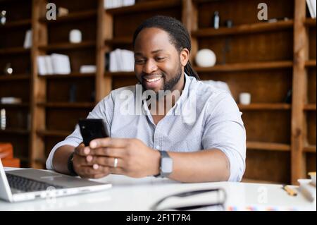 Smiling African-American man wearing smart casual shirt is using smartphone on the workplace at office, a happy mixed-race guy texting messages, enjoying chatting in social networks Stock Photo