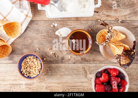 Top view image of a table where hand rolled fresh made waffle cones are dipped into melted chocolate and walnut pieces are sticked on top.  A  bowl of Stock Photo