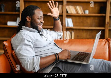 Cheerful African-American man is using a laptop for video communication with friends, family or coworkers, a guy is lying on the sofa, waving hand, greeting online participants, side view Stock Photo