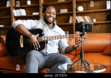 The African-American guy with a guitar is performing online, a biracial smiling man sits on the sofa, play acoustic guitar and streaming online, recording himself on the smartphone, holding a webinar Stock Photo